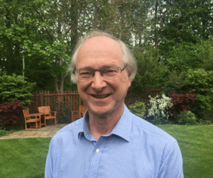 A white balding man in glasses and a blue shirt smiling and standing on a lawn in a wooded area.